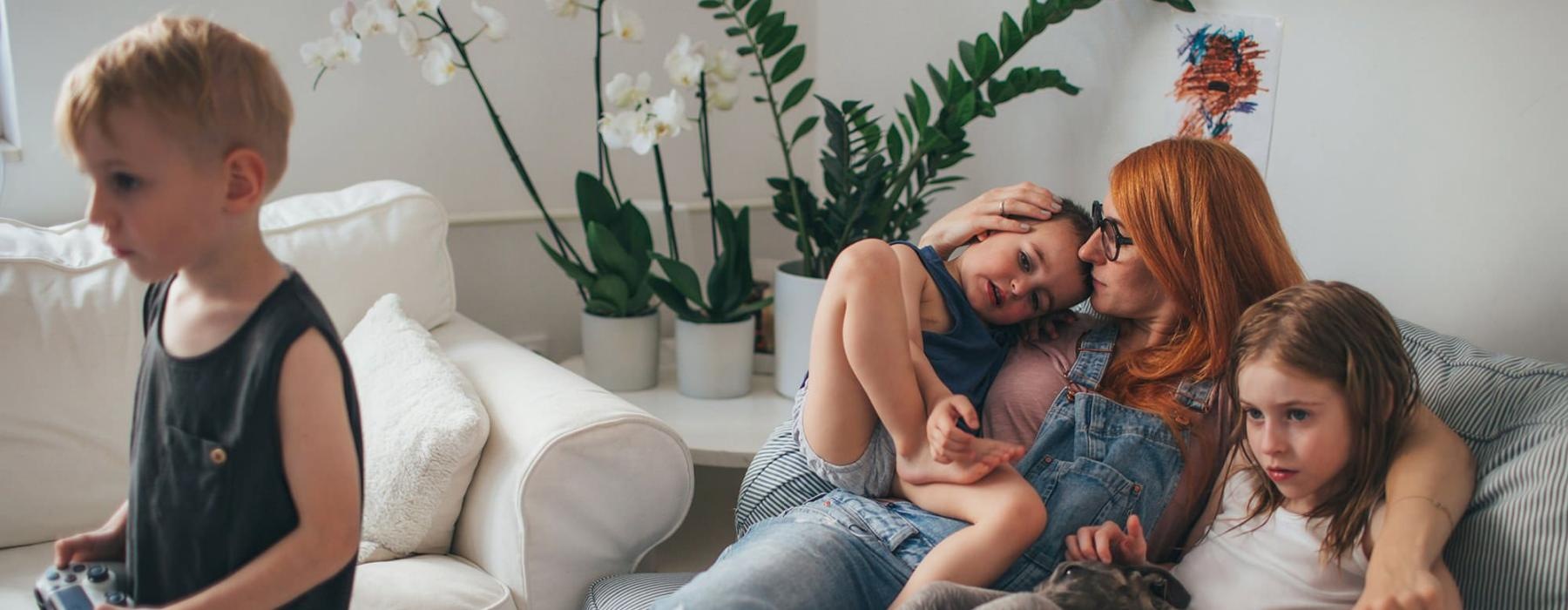 mother sits on a couch with her daughters while her son plays a video game